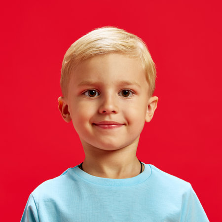 Portrait of adorable little baby boy, blond haired child wearing blue t-shirt and looking at camera with warm smile against red studio backgroundの写真素材