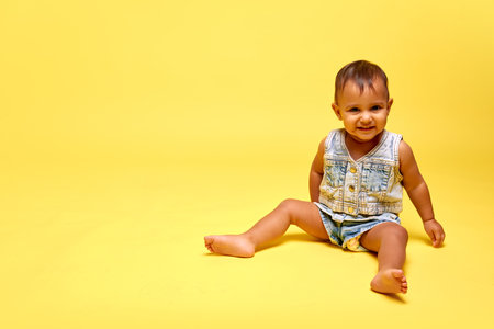 Little baby girl, charming child sitting confidently in denim outfit, smiling against yellow studio backgroundの写真素材