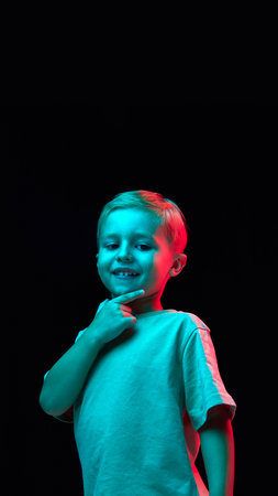 Vertical portrait of cheerful smiling boy, child in t-shirt posing with thoughtful positive face against black studio background in neon lightの写真素材