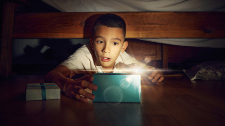 Little emotional boy, child lying on floor in bedroom and looking under bed with flashlight. Excited...