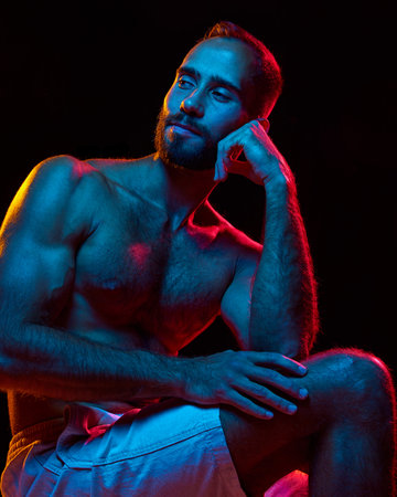 Portrait of handsome bearded young man with muscular shirtless body posing against black studio background in neon lightの写真素材