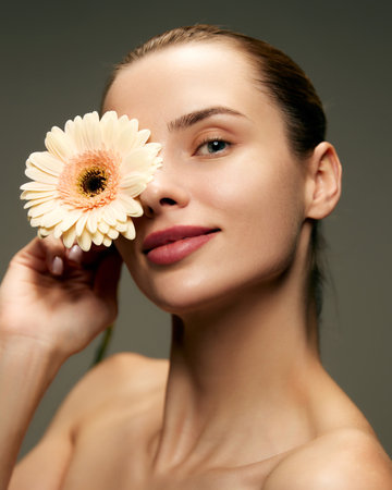 Portrait of beautiful young woman with smooth, spotless skin posing with gerbera flower against green studio backgroundの写真素材