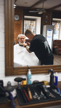 Mirror reflection of man, barber trimming beard of older man sitting in barber chair. Professional grooming environment.の写真素材