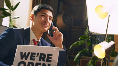 Young businessman sitting at desk in his office and leading conversation via mobile phone with...