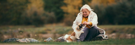 Cozy autumn picnic. Elderly woman sitting on blanket by the river, feeding her dog, spending lovely time with her dog.の写真素材