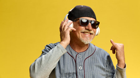 Elderly bearded man in cap and sunglasses listening to music in headphones against yellow studio...