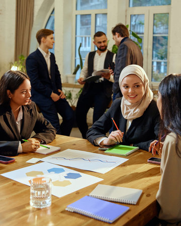 Multicultural female colleagues engaged in discussion during corporate strategy meeting. Other team,...