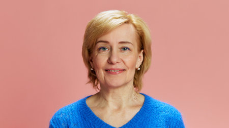 Portrait of elderly, mature woman with blonde short hair, posing in blue sweater, smiling against...