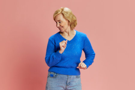 Portrait of happy, smiling, elderly, mature woman in blue sweater, cheerfully dancing, posing against pink studio backgroundの写真素材