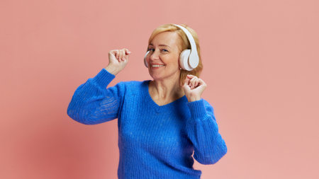 Portrait of happy, smiling, mature woman in blue sweater, listening to music in headphones and dancing against pink studio backgroundの写真素材