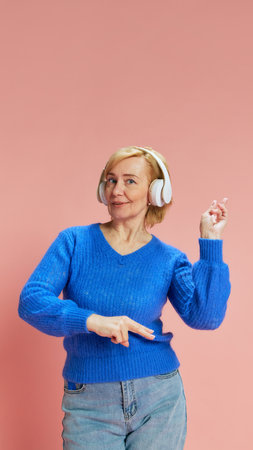 Portrait of happy, smiling, mature woman in blue sweater, listening to music in headphones and dancing against pink studio backgroundの写真素材