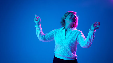 Positive, elderly woman cheerfully dancing, relaxing against blue studio background in neon light