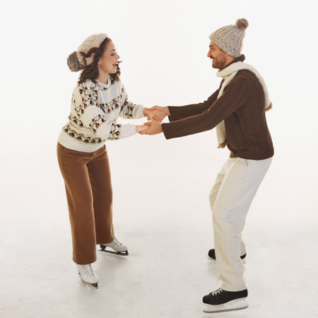 Joyful happy couple, man and woman skating on ice, holding hands, wearing knitted sweaters and hats...