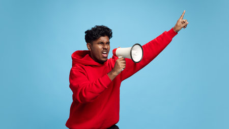Emotional young Indian man shouting in megaphone wand lively gesturing against blue studio...