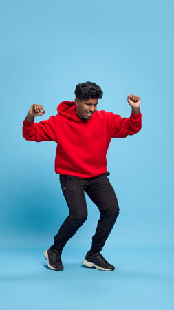 Full-length portrait of cheerful young Indian man in black jeans and red hoodie, cheerful dancing against blue studio background. Positive vibesの写真素材