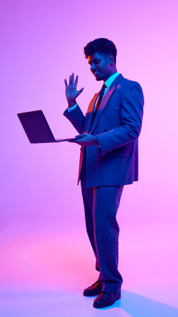 Portrait of young smiling Indian man in formal wear holding laptop and waving into screen, greeting with colleagues against purple studio background in neon lightの写真素材
