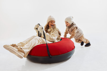 Cozy happy moment between mother and son. Happy mother sitting on red snow tube and little boy pushing it forwardの写真素材