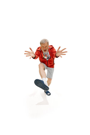 Senior man dressed red jacket, shorts and slippers dancing with joy raising hands against white studio background.の写真素材