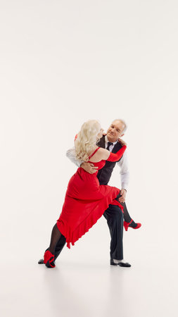 Full-length image of senior couple, man and woman dressed in elegant red dress and black classic suit, dancing tango isolated on white backgroundの写真素材