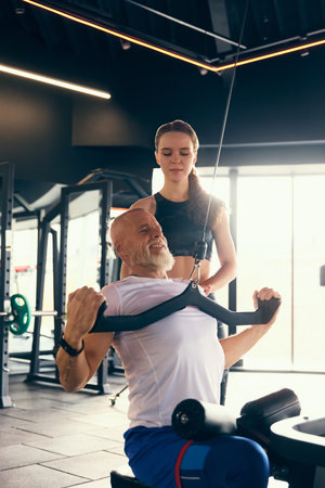 Senior man training, performing lat pulldown exercise, guided by fitness trainer in professional gym, emphasizing back strength.の写真素材