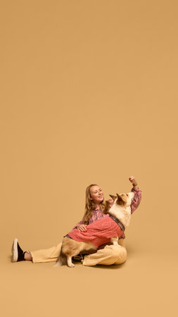 Beautiful young woman in checkered shirt posing with adorable happy Corgi dog wearing pink sweater against warm studio backgroundの写真素材