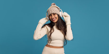 Portrait of happy young Indian woman in knitted hat, wearing headphones and listening to music with...