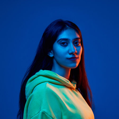 Portrait of young Indian woman wearing yellow hoodie, posing with calm expression against blue studio background in neon lightの写真素材