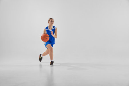 Full-length dynamic image of young teen girl, basketball player in blue uniform in motion with ball during game, training against white studio backgroundの写真素材
