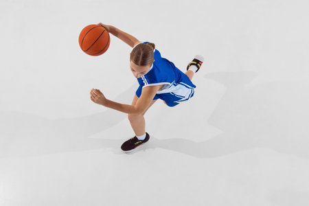 Top view image of competitive teen girl in blue uniform, basketball player during training session, in motion with ball against white studio backgroundの写真素材