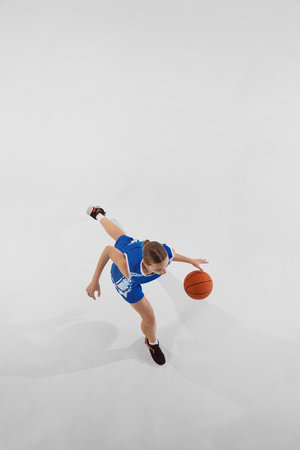 Top view full-length image of teen girl basketball player wearing blue uniform, in motion with ball, training against white studio backgroundの写真素材