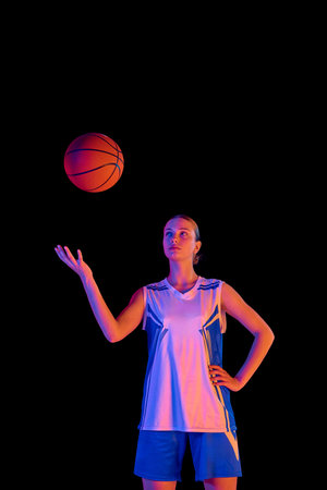 Portrait of young girl in uniform, basketball player bouncing ball on hand against black studio background in neon lightの写真素材