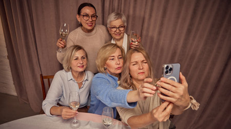 Group of five elderly women, friends, colleagues sitting at home, drinking wine and taking selfie with smartphone. Memorable cozy momentの写真素材