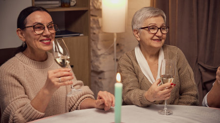 Two women dressed in cozy warm sweaters, sitting at table in warmly lit living room, drinking white wine, talking, smilingの写真素材