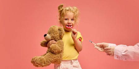 Little girl, child holding teddy bear toy, eating lollipop while doctor making vaccination injection against pink studio backgroundの写真素材