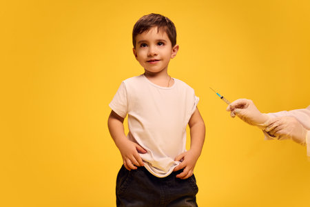 Little boy, child in black and white clothes calmly standing while doctor making vaccine injection with syringe against yellow studio background.の写真素材