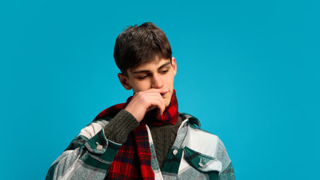 Handsome young man in checkered shirt and red scarf having cold and flu, sneezing against blue studio background. Health care, treatmentの写真素材