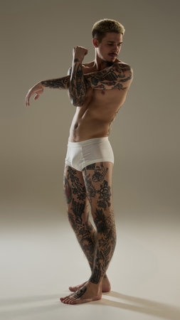 Full-length portrait of handsome young guy with tattoos shirtless body, posing in underwear against neutral studio backgroundの写真素材