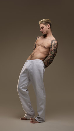 Full-length portrait of handsome young guy with shirtless, athletic, tattooed body design in white sportive pants against neutral studio backgroundの写真素材