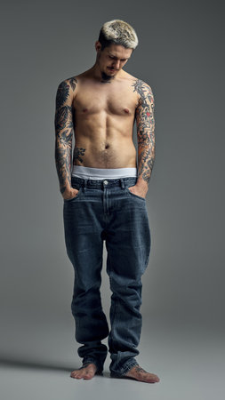 Full-length of handsome young man with blonde hair, muscular tattooed body, posing shirtless in jeans against grey studio backgroundの写真素材
