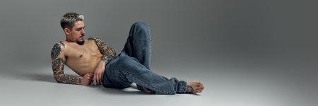 Handsome young man with shirtless muscular tattooed body, sitting on floor, posing in jeans agate grey studio backgroundの写真素材