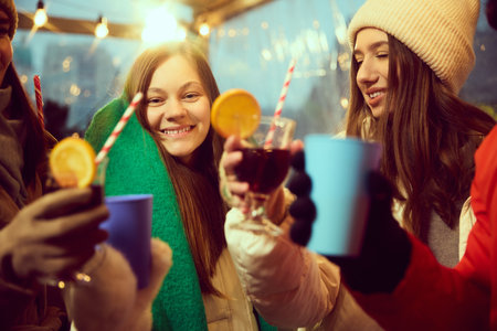 Young smiling women wearing warm hats and scarves drinking hot drinks, mulled wine. Friends gathering at outdoor winter fairの写真素材