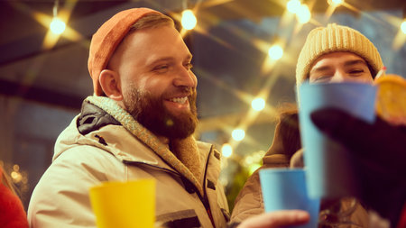 Smiling young bearded man in orange beanie hat meeting with friends, drinking hot drinks, laughing. Cozy moment at outdoor winter eventの写真素材