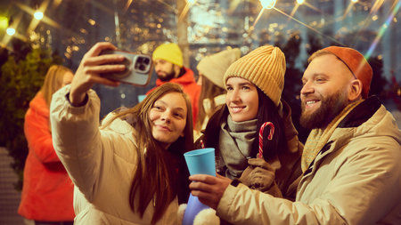 Cheerful group of friends smiling while capturing selfie during winter gathering outdoors, drinking hot drinks, enjoying the festive atmosphere.の写真素材