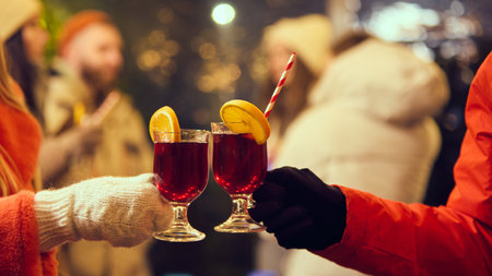 Two glasses of mulled wine with orange slices and striped straws clinking together, celebrating a cozy winter evening under festive lights.の写真素材