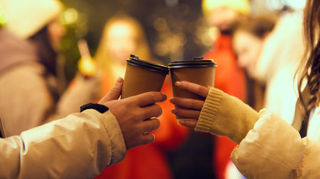 Close-up of two disposable coffee cups being toasted together at cozy winter gathering with blurred...