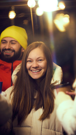 Young happy woman in white jacket smiling brightly at festive winter fair, surrounded by string lights and warm holiday vibes.の写真素材
