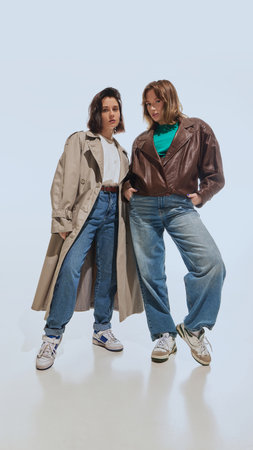 Full-length portrait of beautiful young women, blonde and brunette, wearing trench coat and leather jacket with denim jeans, posing against white background.の写真素材