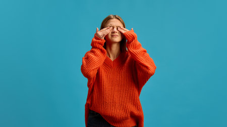 Young woman in orange sweater closing her eyes appearing in discomfort isolated on a blue...
