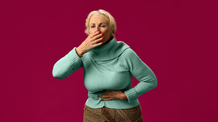 Elderly woman coughing while holding stomach, experiencing abdominal discomfort from excessive coughing on pink studio backgroundの写真素材