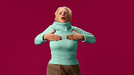 Senior woman holding hands on chest, suffering from pain, asthma attack on pink studio background. Difficulty in breathing and chest tightness.の写真素材
